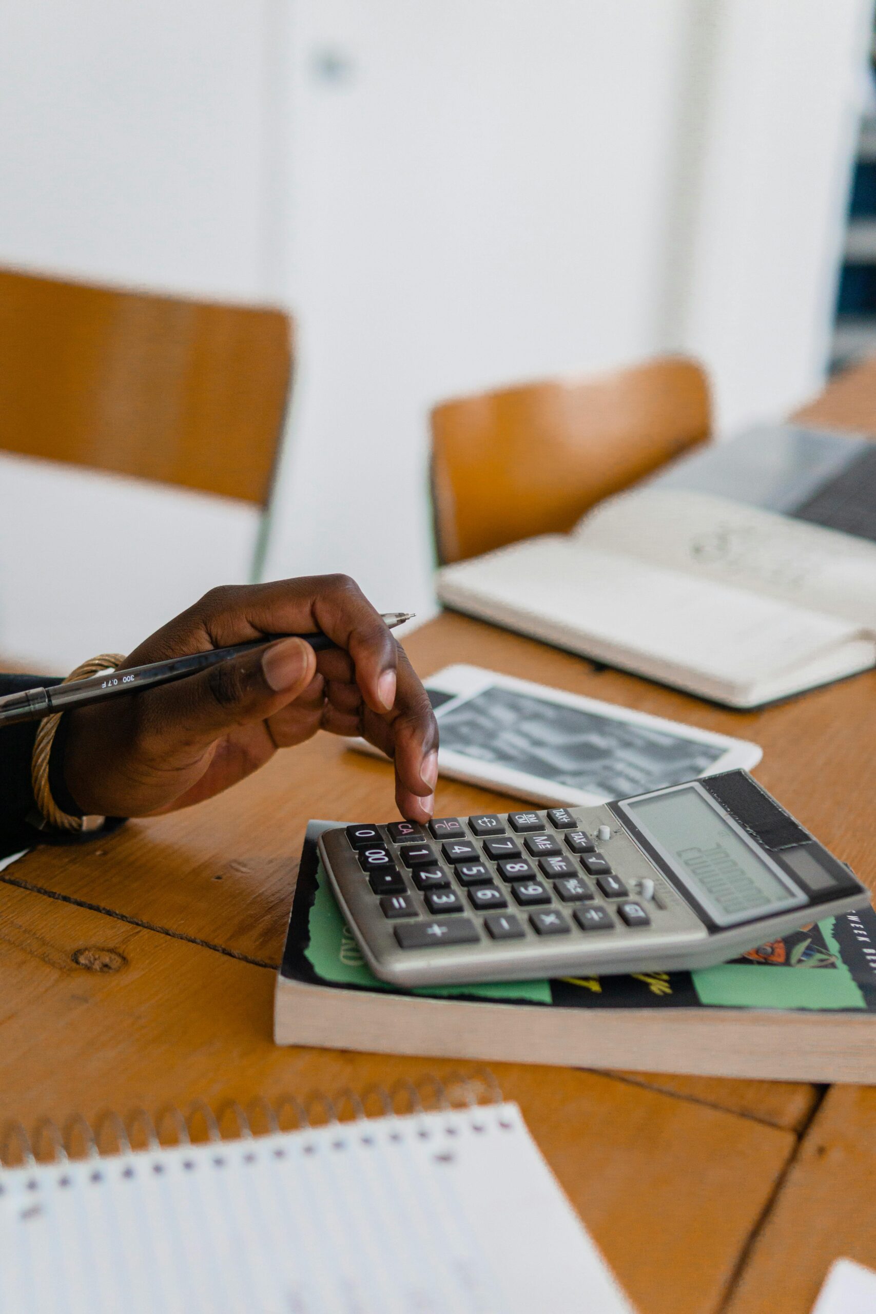 A person using a calculator on a wooden table, surrounded by books and stationery.